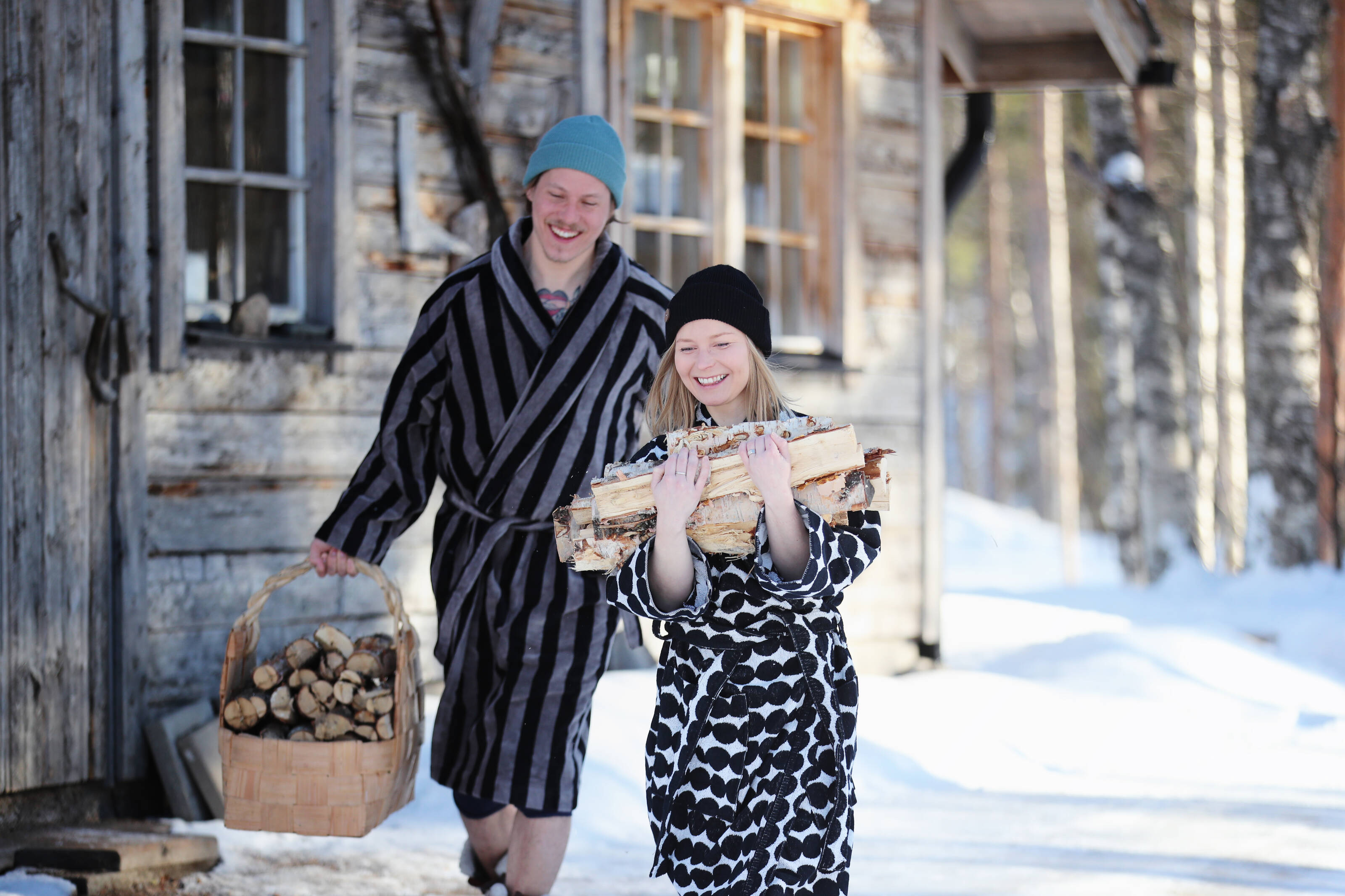 Two smiling people carrying wood outside a Finnish sauna